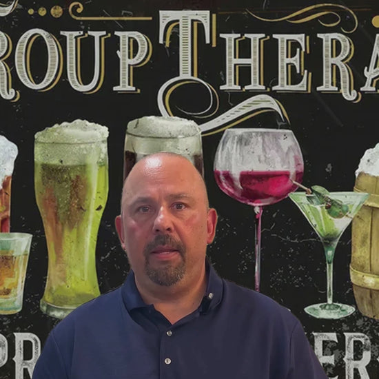 Promotional video for Group Therapy Practiced Here metal tin sign showing presenter in navy polo shirt standing in front of sign featuring hand-drawn illustrations of beer glasses, wine glass, and cocktails with GROUP THERAPY typography in cream lettering on dark brown background with decorative scrollwork