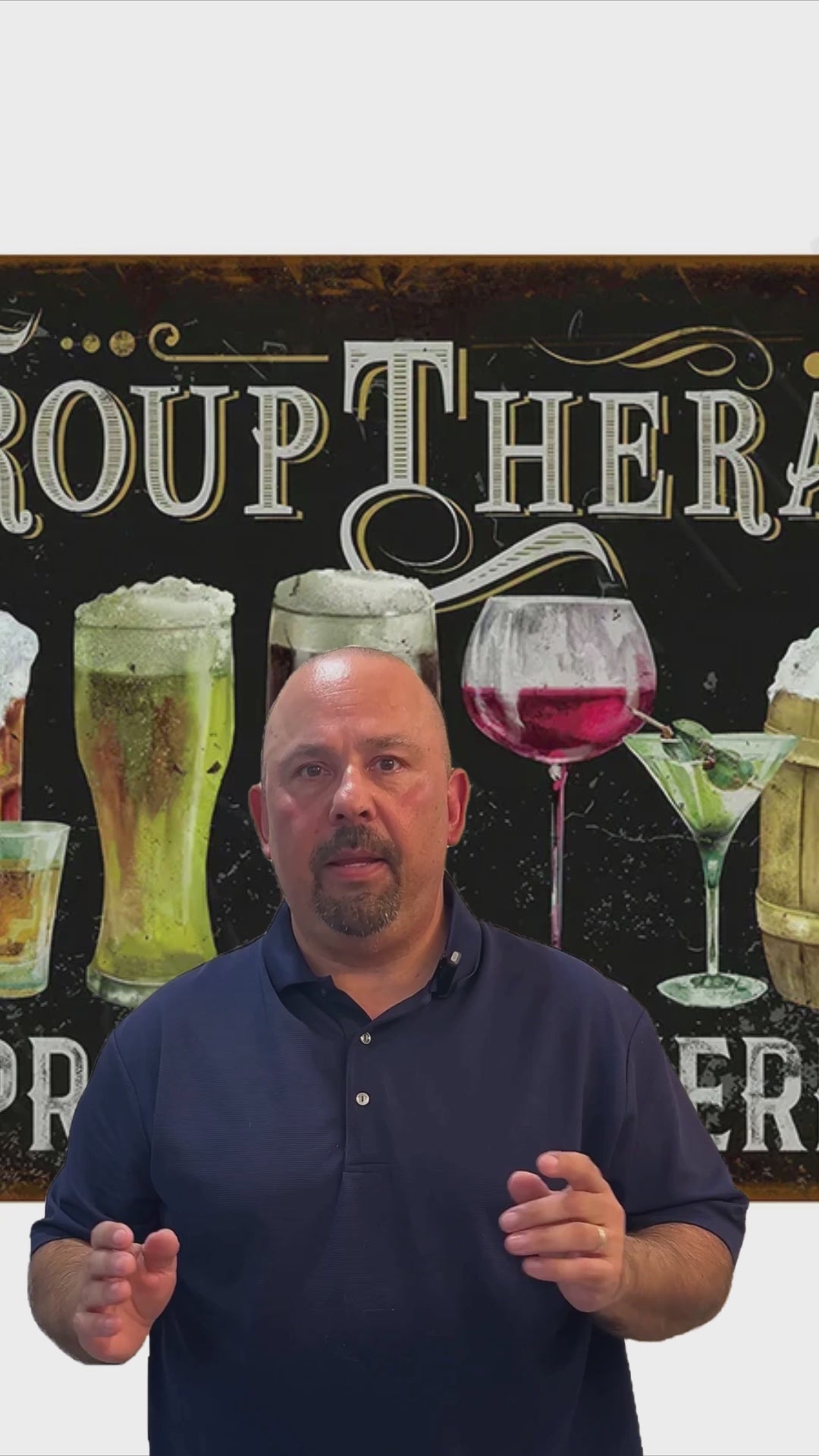 Promotional video for Group Therapy Practiced Here metal tin sign showing presenter in navy polo shirt standing in front of sign featuring hand-drawn illustrations of beer glasses, wine glass, and cocktails with GROUP THERAPY typography in cream lettering on dark brown background with decorative scrollwork